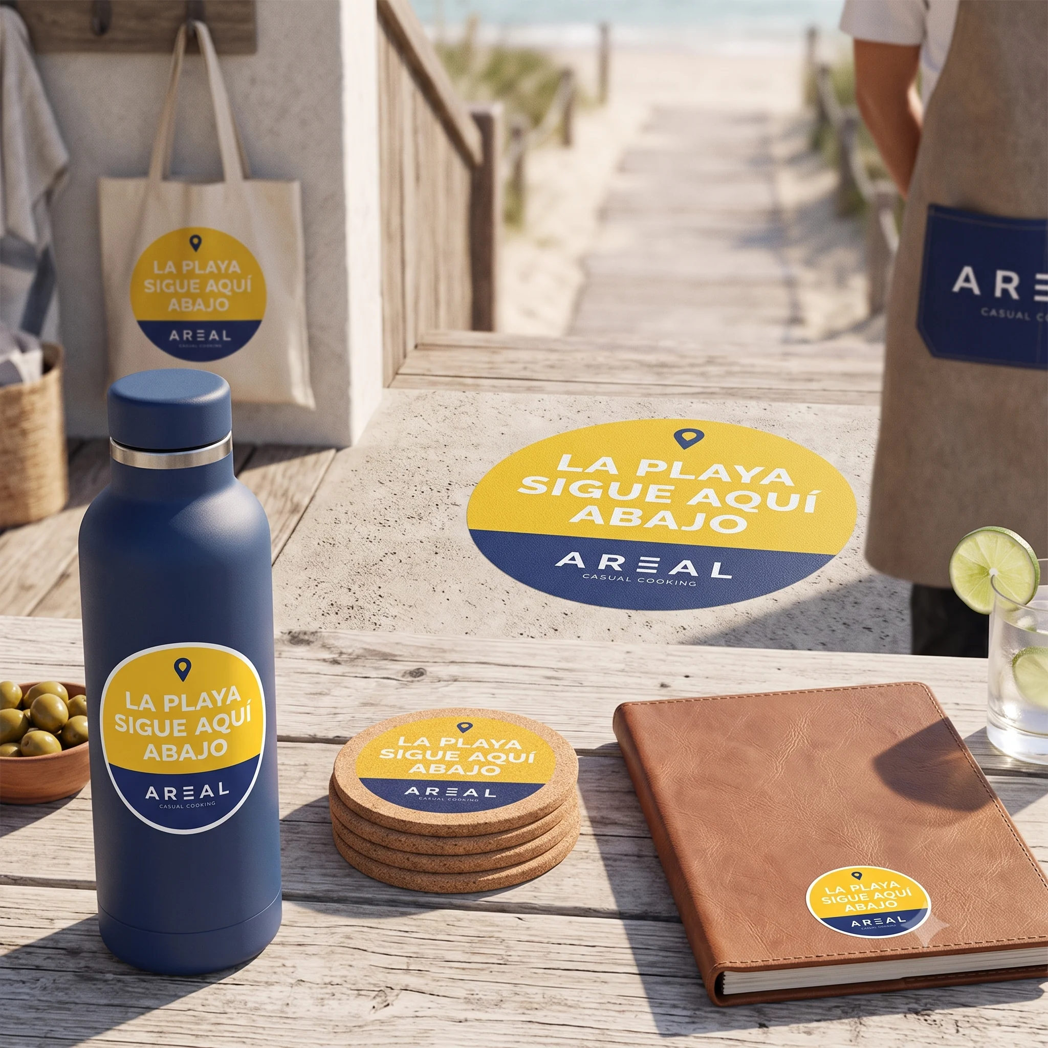 Sello circular AREAL con el claim la playa sigue aquí abajo aplicado como pegatina en una bolsa de tela, botella, posavasos de corcho, libreta y bolsillo de delantal, en un entorno de playa